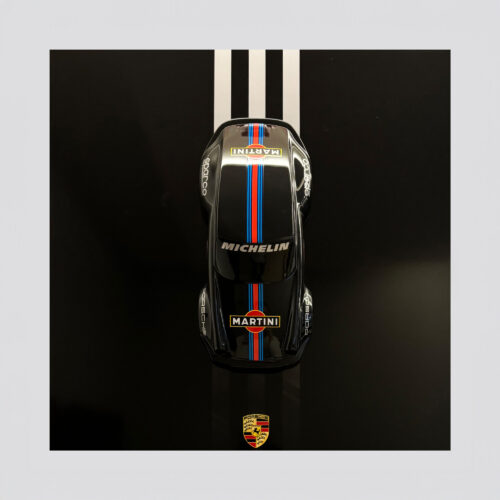 Top view of a racing car toy with Martina and Michelin logos, positioned on a black surface with three white lines, and a Porsche logo at the bottom.