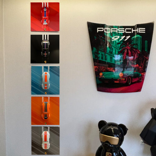 Wall displaying five art pieces of miniature Porsche cars in different colors next to a large Porsche 911 banner and a black bear statue wearing sunglasses.