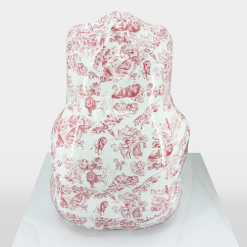 A sculptural object with a rounded, abstract shape covered in red toile de Jouy patterns depicting various animals and nature scenes on a white background, placed on a reflective surface.