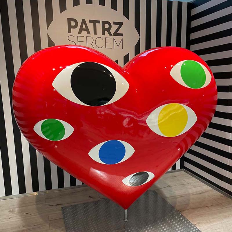 Large red heart sculpture with multiple colorful eyes on it, against a black and white striped background with Polish text 