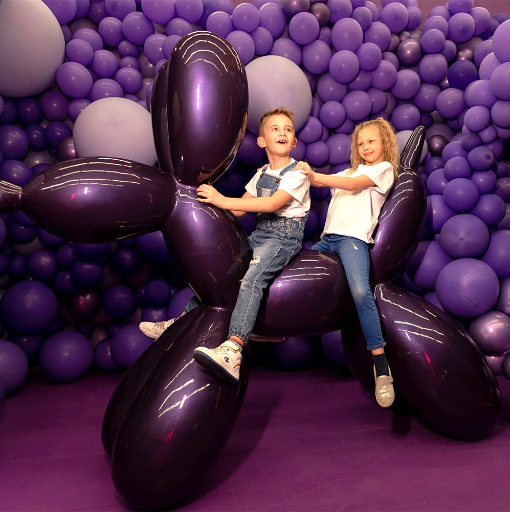Large Decorative Figure Balloon Dog - Metallic Purple 3 Two children playing on a large, dark purple balloon sculpture of a dog, surrounded by various sized purple balloons.