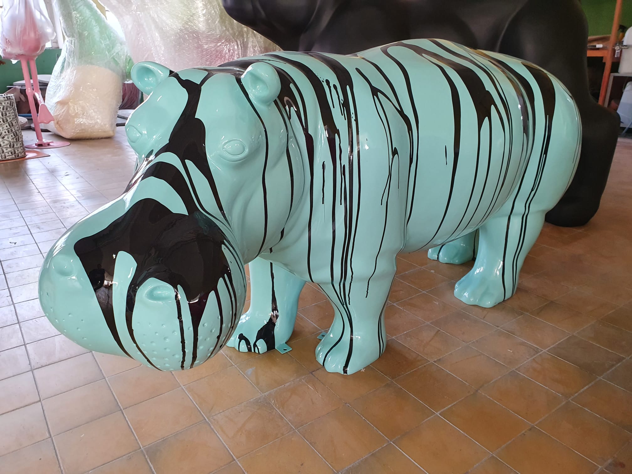 3D Figure Made of Laminate - Life-Size Hippopotamus - Trash 3 A turquoise hippopotamus statue with black paint streaks on its body, standing on a tiled floor in an indoor setting with wrapped objects in the background.