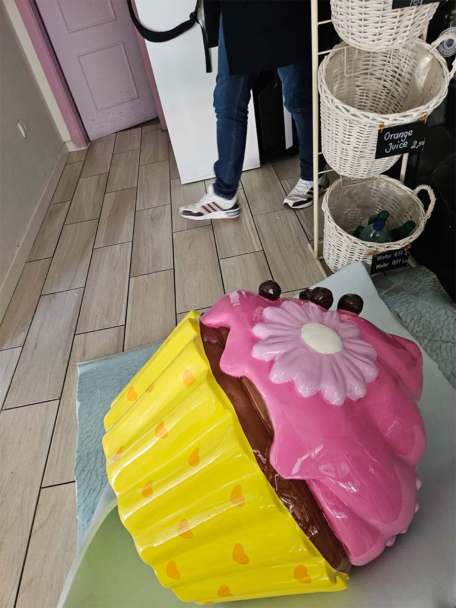 Colorful Cupcake for Sweet Home Bakery in Lagos, Portugal 3 Rzeźba wielkiego babeczki w pastelowych kolorach stojąca na podłodze w kawiarni obok osoby w dżinsach i białych butach.