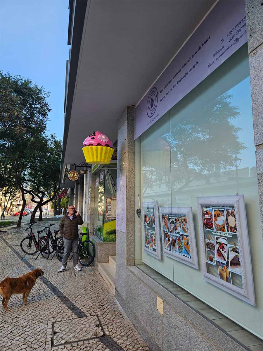 oryg_064490685_1129949550544944_1465385417682452480_o Street scene with a man standing near bicycles and talking on the phone outside a bakery shop. A brown dog is standing nearby. The bakery sign features a large cupcake, and the shop window displays pictures of desserts.