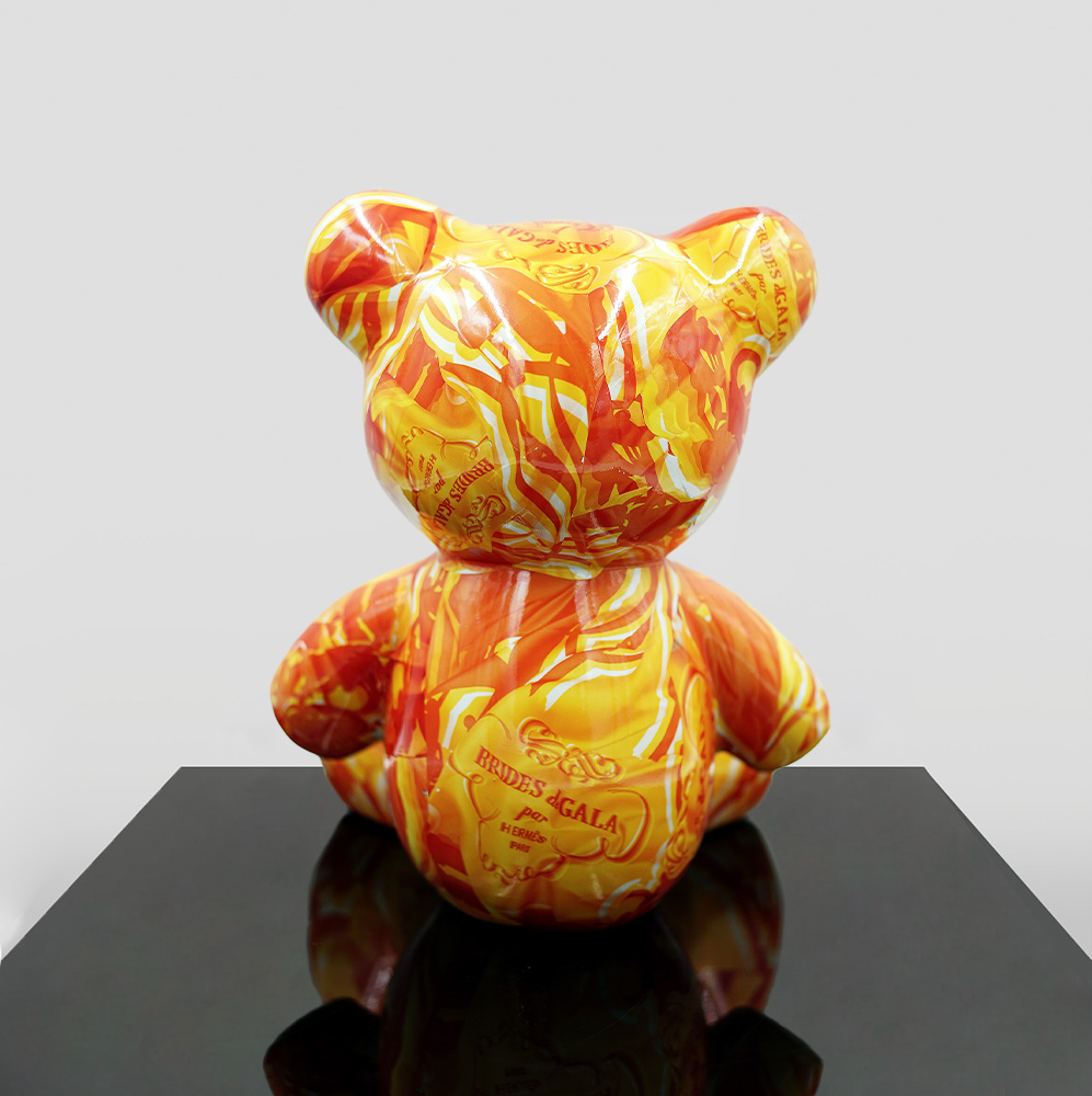 Small Sitting Bear - Orange H 2 A colorful bear-shaped sculpture with an orange and yellow swirling pattern sits on a black reflective surface.