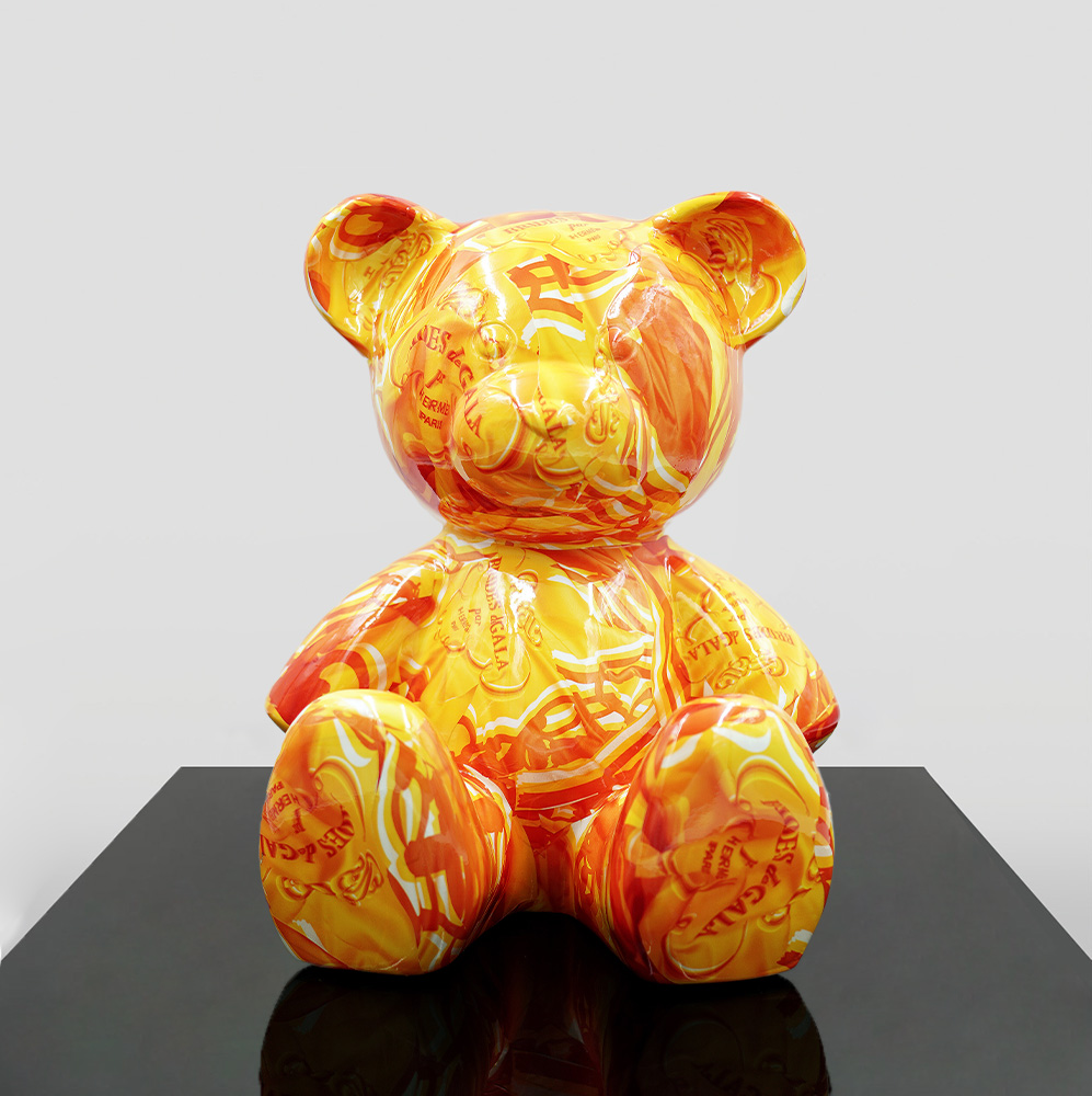 Small Sitting Bear - Orange H 1 A large, stylized teddy bear sculpture with a vibrant orange and yellow pattern featuring various text and images. The sculpture sits on a glossy black surface against a plain white background.