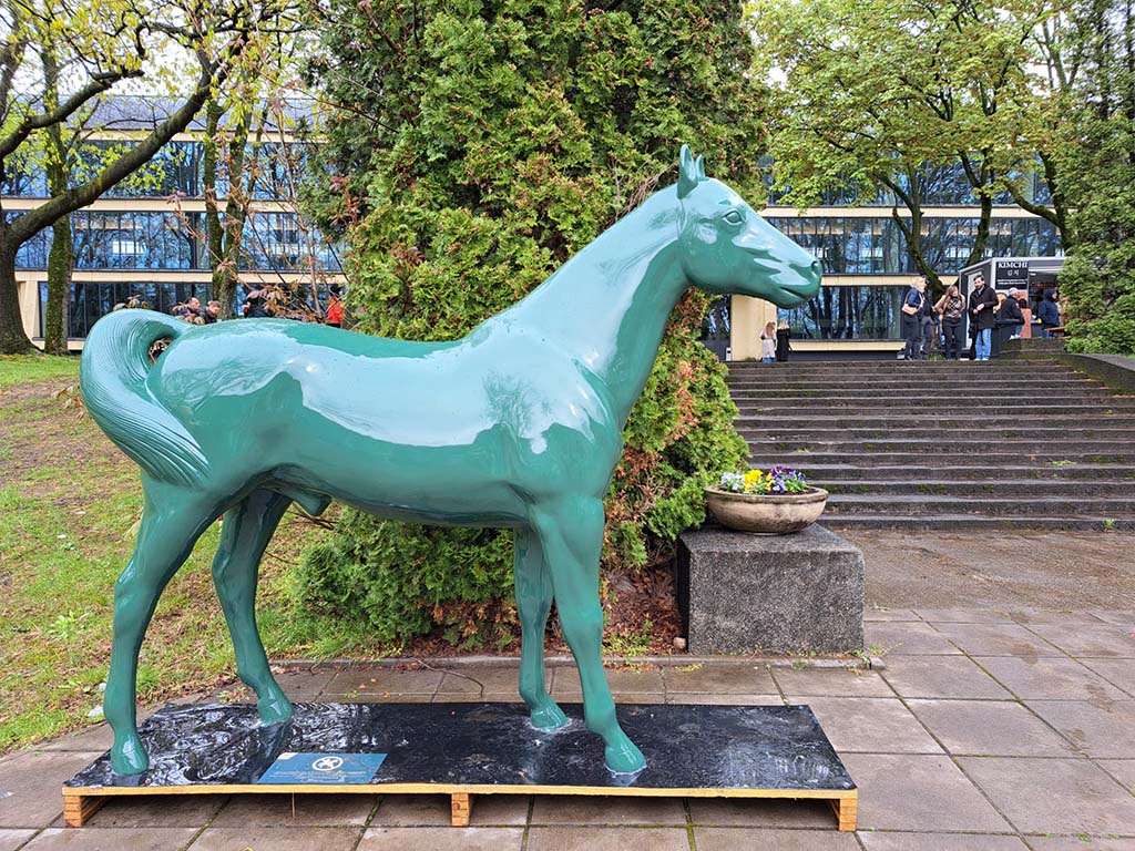 Green horse statue on a wooden platform, placed on paved ground, surrounded by trees and a building with steps in the background.