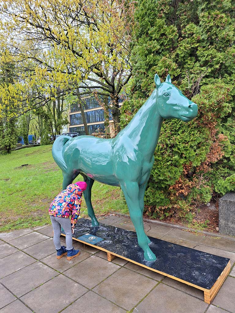 Dziecko w kolorowej kurtce zaglądające pod rzeźbę zielonego konia na zewnątrz w parku.