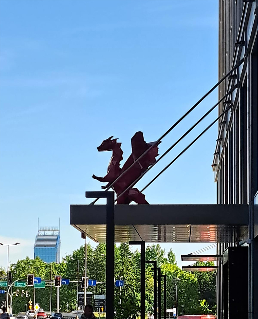 Low Poly Dragons at the Radisson Hotel 6 Czerwony smok na daszku budynku, w tle miejski krajobraz z drzewami i budynkami.