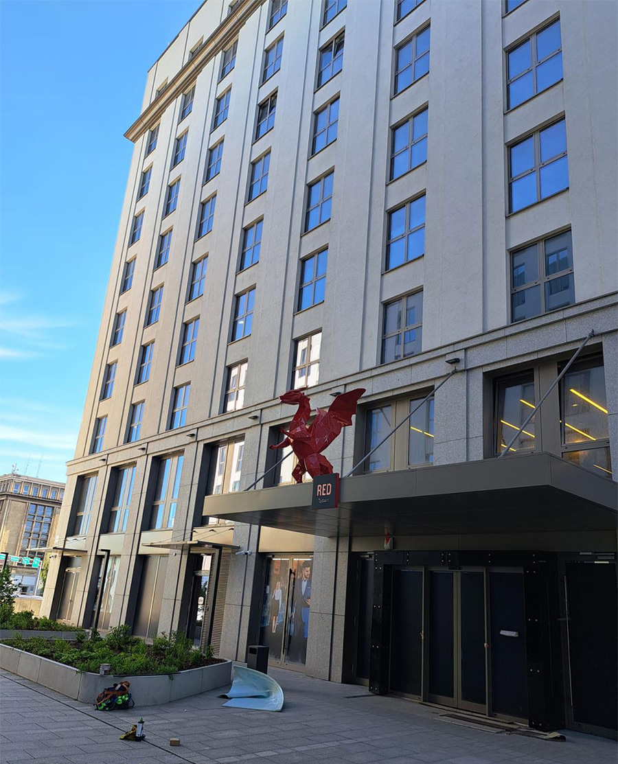 Low Poly Dragons at the Radisson Hotel 8 Wysoki budynek z szarymi ścianami i licznymi oknami, przed wejściem czerwony posąg smoka z napisem