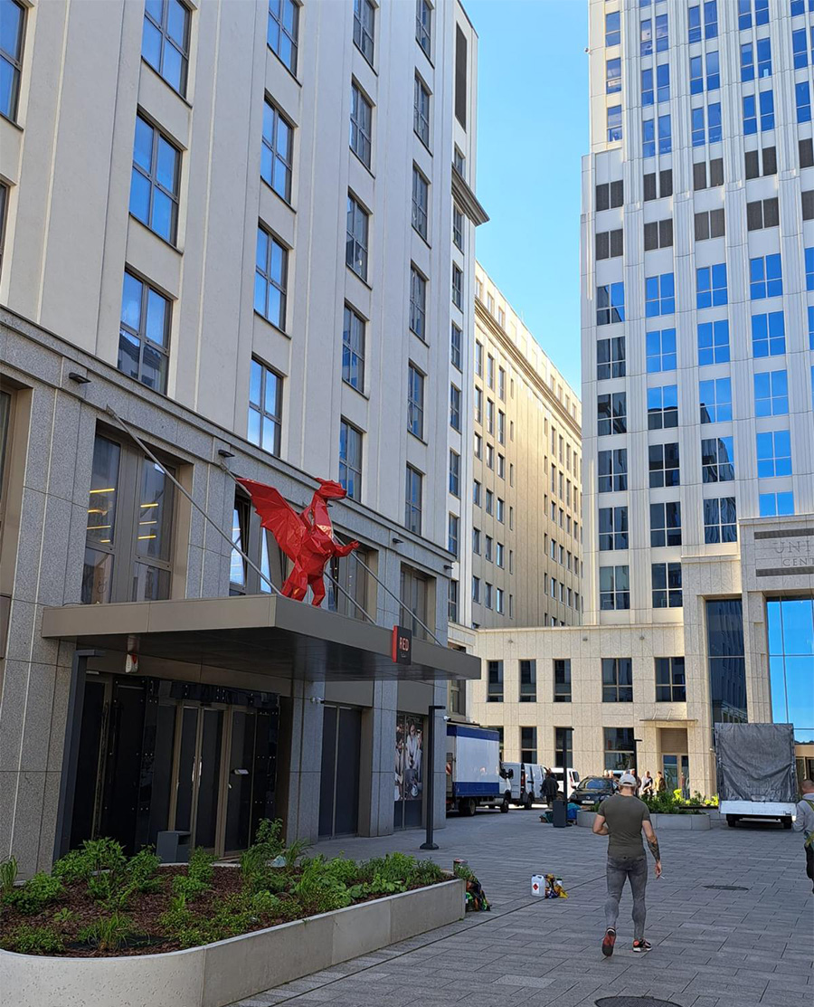 Low Poly Dragons at the Radisson Hotel 5 Budynek z czerwoną rzeźbą konia na daszku oraz osoba przechodząca chodnikiem.