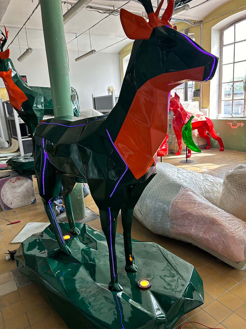 Jägermeister Deer with Lighting 8 Duży, kolorowy posąg jelenia wewnątrz budynku, otoczony innymi podobnymi rzeźbami i pakunkami.