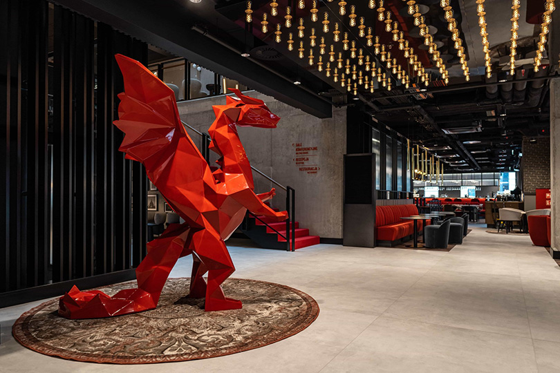 Low Poly Dragons at the Radisson Hotel 2 Czerwona, geometryczna rzeźba smoka w nowoczesnym wnętrzu hotelu, z dużym dywanem i stylowym oświetleniem.