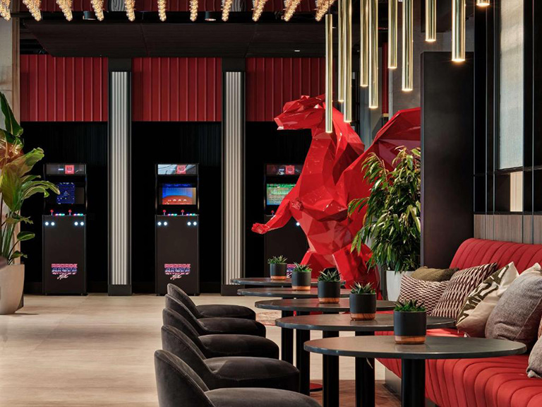 Low Poly Dragons at the Radisson Hotel 3 Wnętrze nowoczesnego salonu z czerwoną ławą, czarnymi fotelami, stylowymi stołami i dużą czerwoną rzeźbą smoka, w tle automaty do gier retro.