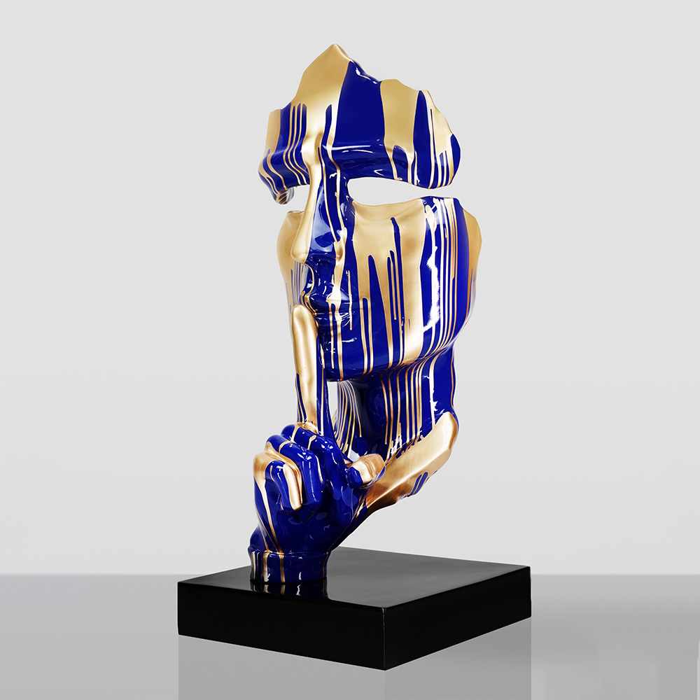 Duża maska na podstawie – w niebieskie łaty 3 Abstract sculpture of a fragmented human face in blue and gold stripes with a hand holding a finger to the lips in a shushing gesture, mounted on a black rectangular base against a plain light gray background.