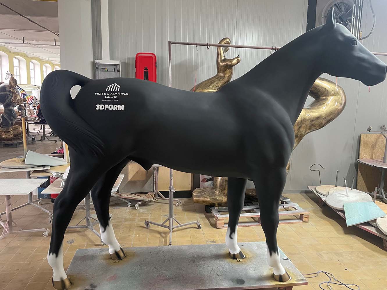 Czarny model konia z białymi kopytami i napisami hotelu Marina Club oraz 3DFORM na boku, stojący w warsztacie rzeźbiarskim z innymi rzeźbami w tle.