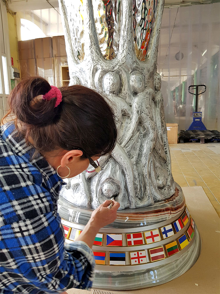 UEFA Europa League Trophy Figure Displayed in Gdańsk During the 2021 Championship 3 Kobieta w niebiesko-czarnej koszuli w kratę pracuje nad dużą srebrną rzeźbą z motywem ludzkich sylwetek, umieszczoną na podstawie ozdobionej flagami różnych państw.