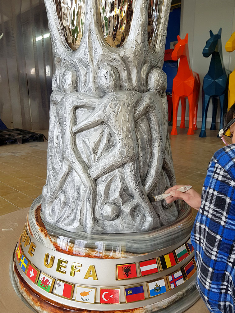 UEFA Europa League Trophy Figure Displayed in Gdańsk During the 2021 Championship 4 Postać malująca rzeźbę przedstawiającą ludzi grających w piłkę nożną na podstawie z flagami krajów i napisem EUROPE UEFA, we wnętrzu z kolorowymi rzeźbami koni w tle