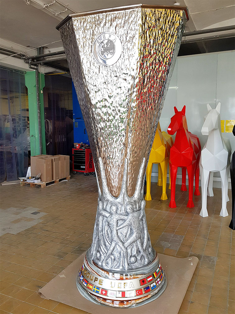 UEFA Europa League Trophy Figure Displayed in Gdańsk During the 2021 Championship 5 Duży srebrny puchar piłkarski z wygrawerowanymi postaciami ludzkimi u podstawy i napisem COUPE UEFA z flagami państw na dolnej części, stojący na podłodze w pomieszczeniu z kolorowymi rzeźbami koni w tle.
