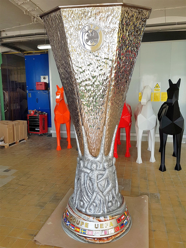 UEFA Europa League Trophy Figure Displayed in Gdańsk During the 2021 Championship 6 Duży srebrny puchar z napisem COUPE UEFA na podstawie, stojący w pomieszczeniu z pomalowaną podłogą, w tle kolorowe geometryczne rzeźby koni w kolorach czerwonym, białym i czarnym oraz narzędzia i kartony.