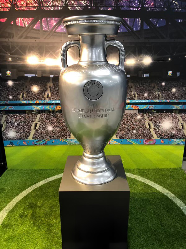 Srebrny puchar UEFA European Football Championship ustawiony na czarnym podium na tle stadionu piłkarskiego z publicznością i oświetleniem.