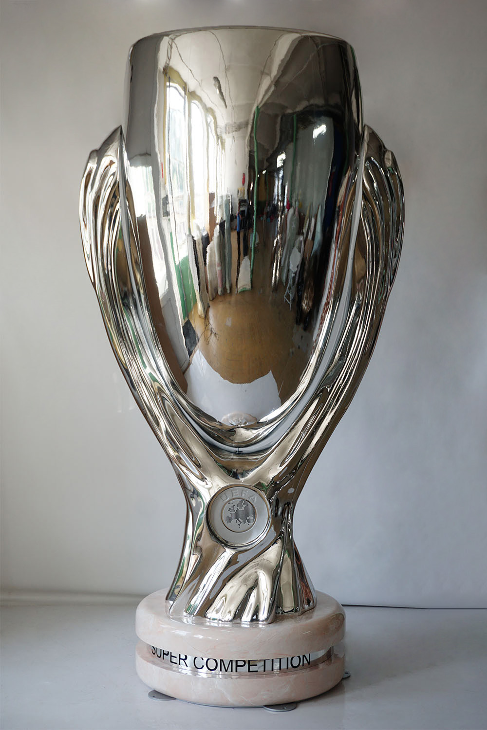 Large UEFA Super Cup Figure for the 2024 Championship 3 Połyskujący srebrny puchar UEFA Super Competition stojący na jasnym marmurowym podstawkach na białym tle.