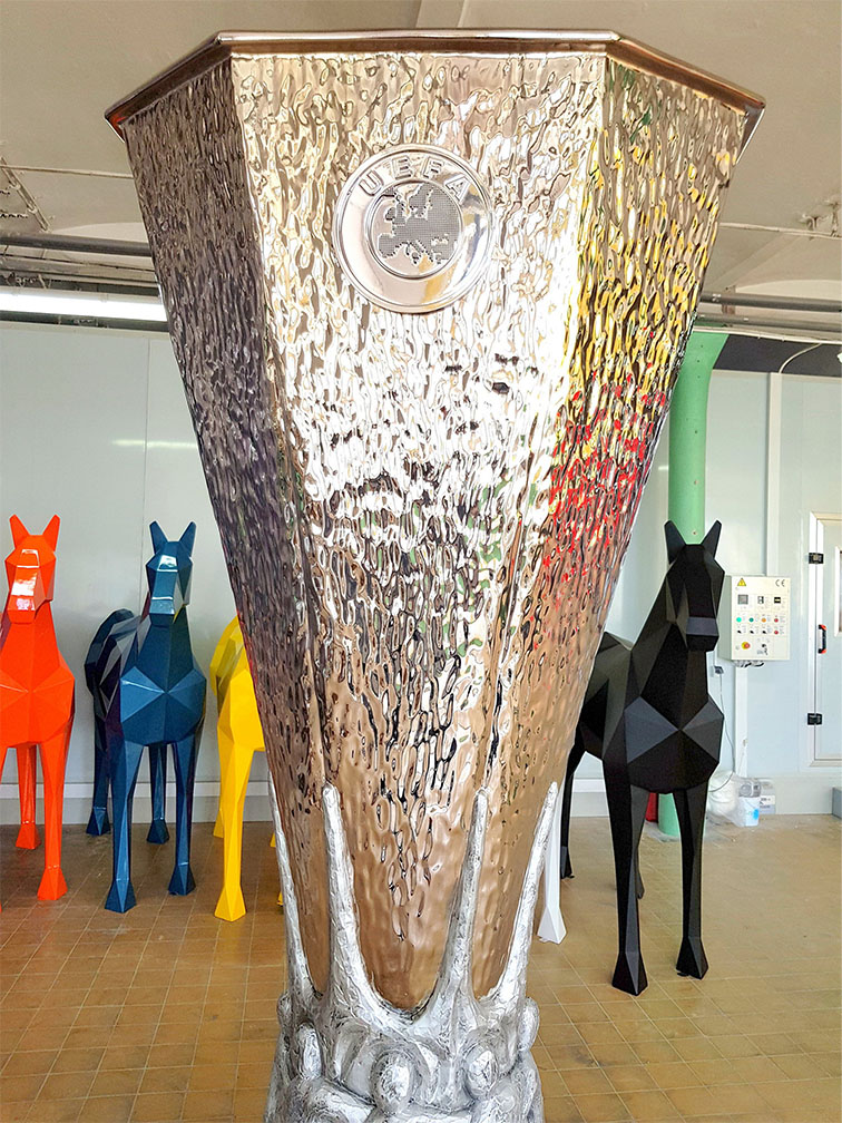 UEFA Europa League Trophy Figure Displayed in Gdańsk During the 2021 Championship 1 Duży srebrny puchar UEFA stojący na podłodze w pomieszczeniu z mozaikową podłogą, w tle kolorowe geometryczne rzeźby koni w kolorach pomarańczowym, niebieskim, żółtym i czarnym.