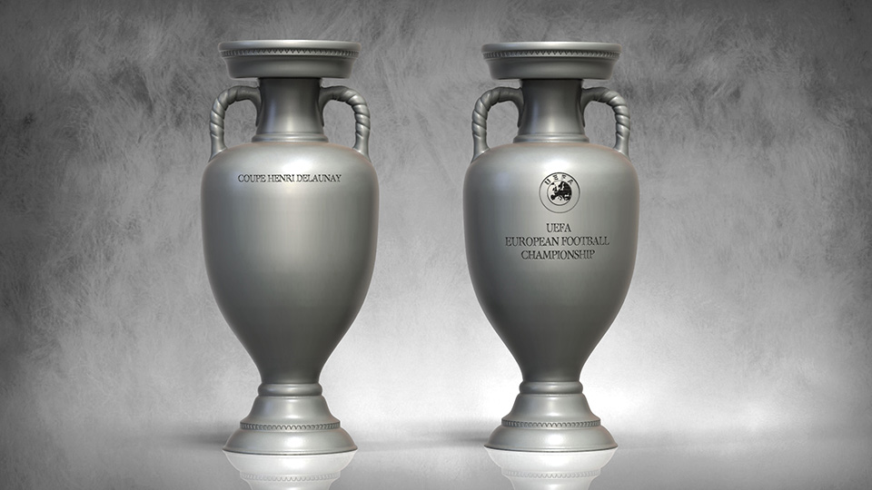 Dwa srebrne puchary UEFA European Football Championship stojące obok siebie na jasnym tle, jeden z napisem COUPE HENRI DELAUNAY, drugi z logo UEFA i napisem UEFA EUROPEAN FOOTBALL CHAMPIONSHIP.