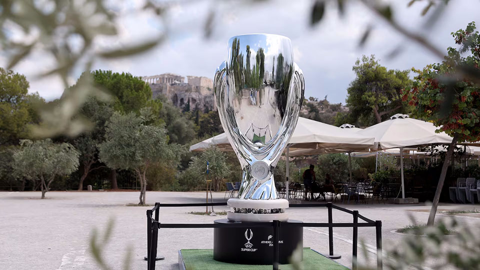 Large UEFA Super Cup Figure for the 2024 Championship 1 Srebrny puchar UEFA Super Cup ustawiony na czarnym podstawie z napisem, umieszczony na otwartej przestrzeni na tle zalesionego terenu i historycznych ruin na wzgórzu.