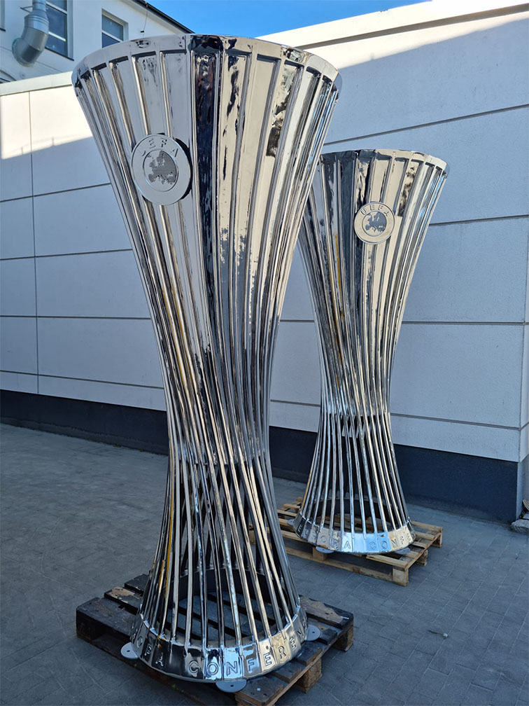 Dwa duże srebrne trofea Europa Conference League ustawione na drewnianych paletach przed jasnym budynkiem na zewnątrz, na szarej nawierzchni.