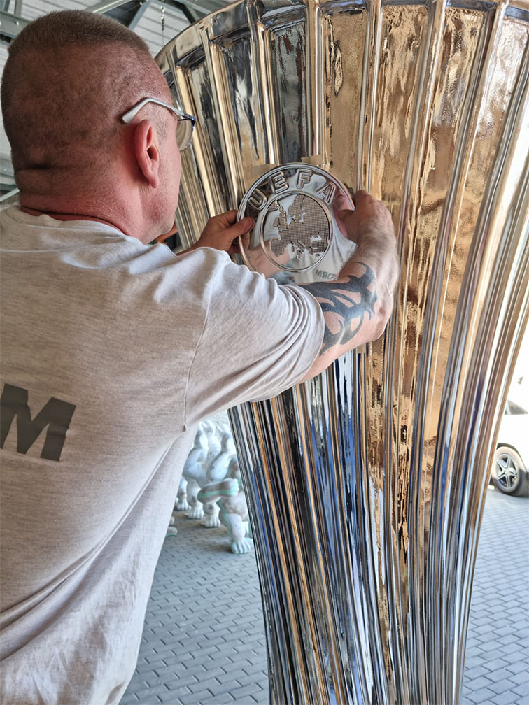 Mężczyzna w szarej koszulce z tatuażem na ramieniu zamocowujący metalowy emblemat UEFA na dużym, srebrnym trofeum.