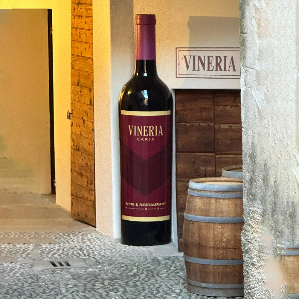 Classic XL Wine Bottle 2 Large decorative wine bottle with a label reading 'Vineria Carin Wine & Restaurant' standing outside a building next to a wooden door and two wooden wine barrels on a cobblestone street.