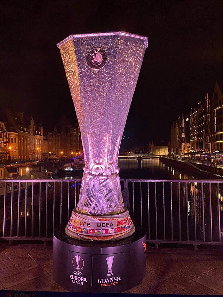 UEFA Europa League Trophy Figure Displayed in Gdańsk During the 2021 Championship 2 Replika trofeum UEFA Europa League na czarnym postumencie z napisem Gdańsk Final 2021, ustawiona na tle nocnej panoramy miasta z oświetlonymi zabytkowymi budynkami i wodą w tle