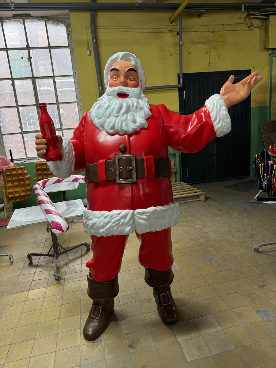 Anyone Can Be Santa: Coca-Cola's Christmas Campaign with Santa Figures 17 Figurka Świętego Mikołaja w czerwonym stroju i brązowych butach stojąca na podłodze w pomieszczeniu z żółto-zielonymi ścianami i dużym oknem, trzymająca czerwoną butelkę w jednej ręce i gestykulująca drugą.