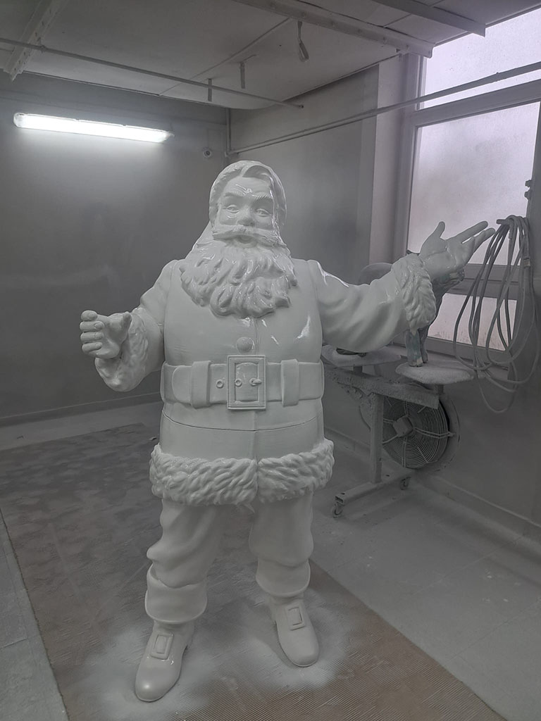 Anyone Can Be Santa: Coca-Cola's Christmas Campaign with Santa Figures 18 Biały, niepomalowany posąg Świętego Mikołaja w pracowni malarskiej, z uniesionymi rękoma, obok okno i wentylator.