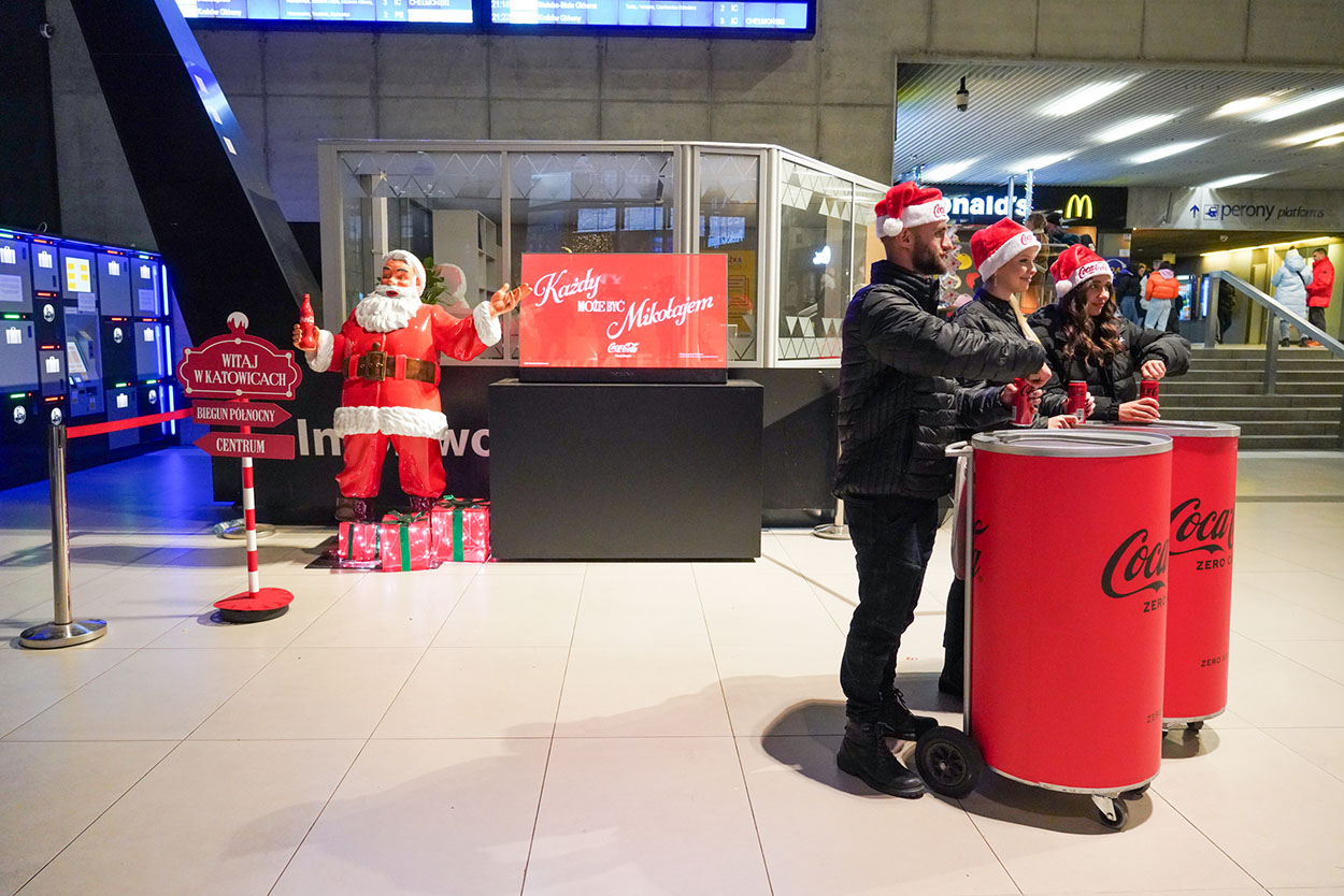 Anyone Can Be Santa: Coca-Cola's Christmas Campaign with Santa Figures 3 Trzy osoby w czapkach św. Mikołaja stoją przy czerwonych pojemnikach Coca-Coli w hali dworca, obok stoi figura św. Mikołaja z butelką Coca-Coli i znakiem