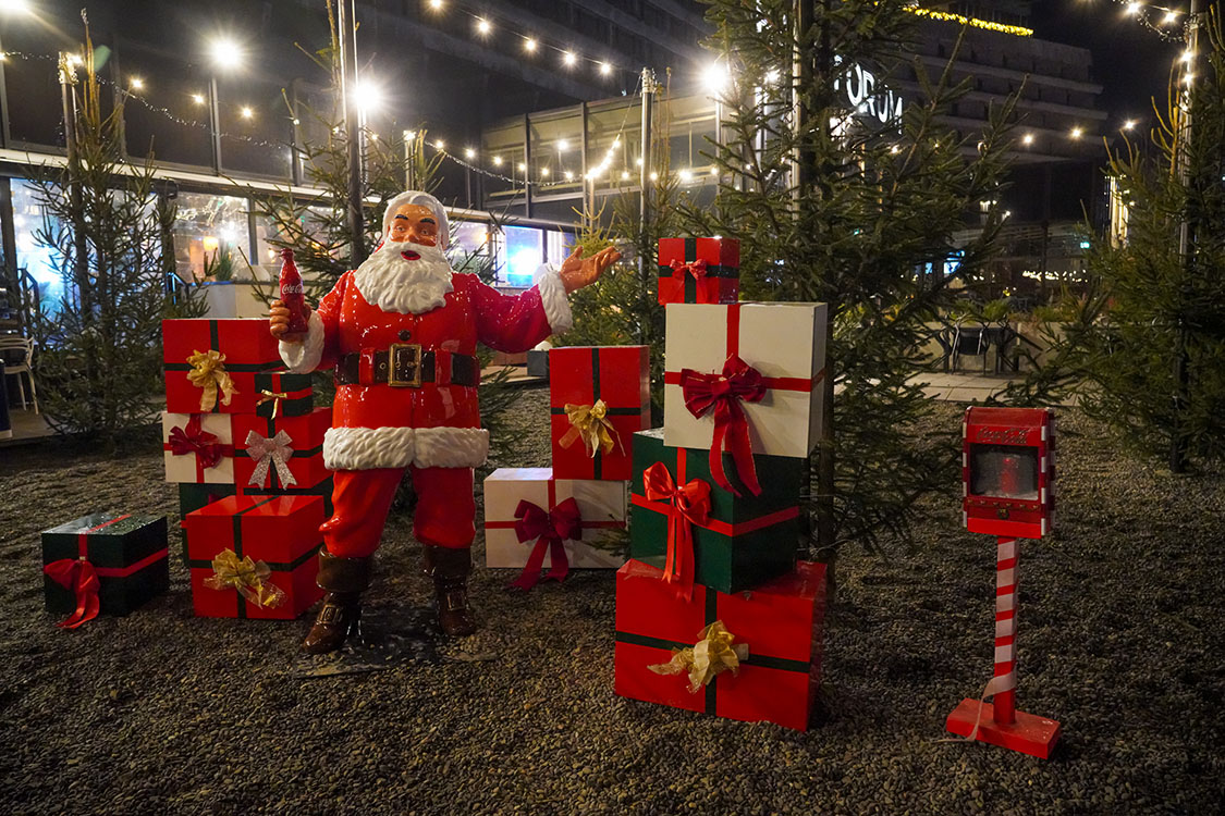Anyone Can Be Santa: Coca-Cola's Christmas Campaign with Santa Figures 1 Figurka Świętego Mikołaja trzymającego butelkę Coca-Coli, otoczona licznymi prezentami w czerwonych, białych i zielonych opakowaniach z kokardami, ustawiona na żwirowym podłożu na tle drzewek iglastych i oświetlenia świątecznego nocą