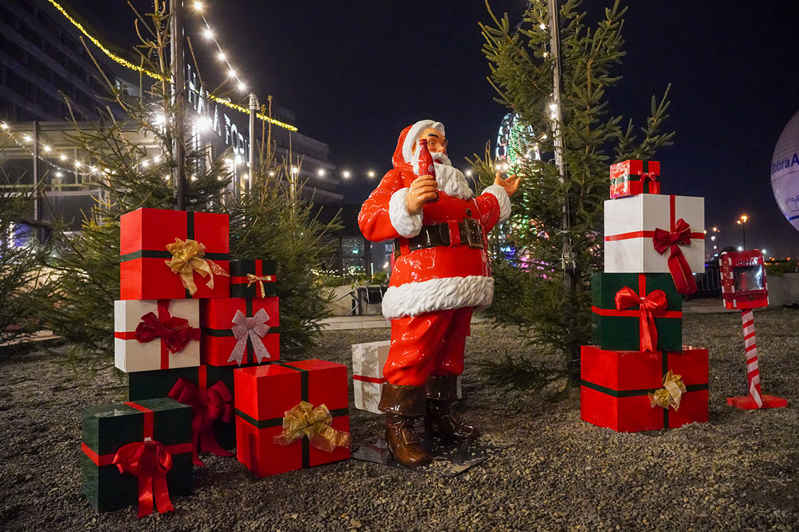 Anyone Can Be Santa: Coca-Cola's Christmas Campaign with Santa Figures 2 Figurka Mikołaja trzymającego butelkę napoju w otoczeniu świątecznie udekorowanych prezentów i małych drzewek iglastych oświetlonych lampkami nocą w plenerze.