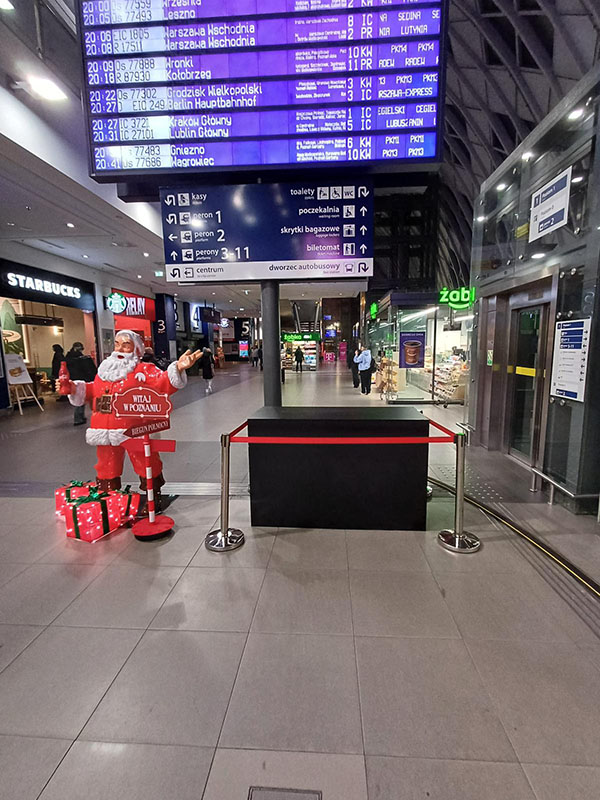Anyone Can Be Santa: Coca-Cola's Christmas Campaign with Santa Figures 7 Wnętrze dworca z tablicą odjazdów pociągów na górze, na dole figura Mikołaja z prezentami i napisem Witaj w Poznaniu oraz sklepami Starbucks i Żabka w tle.