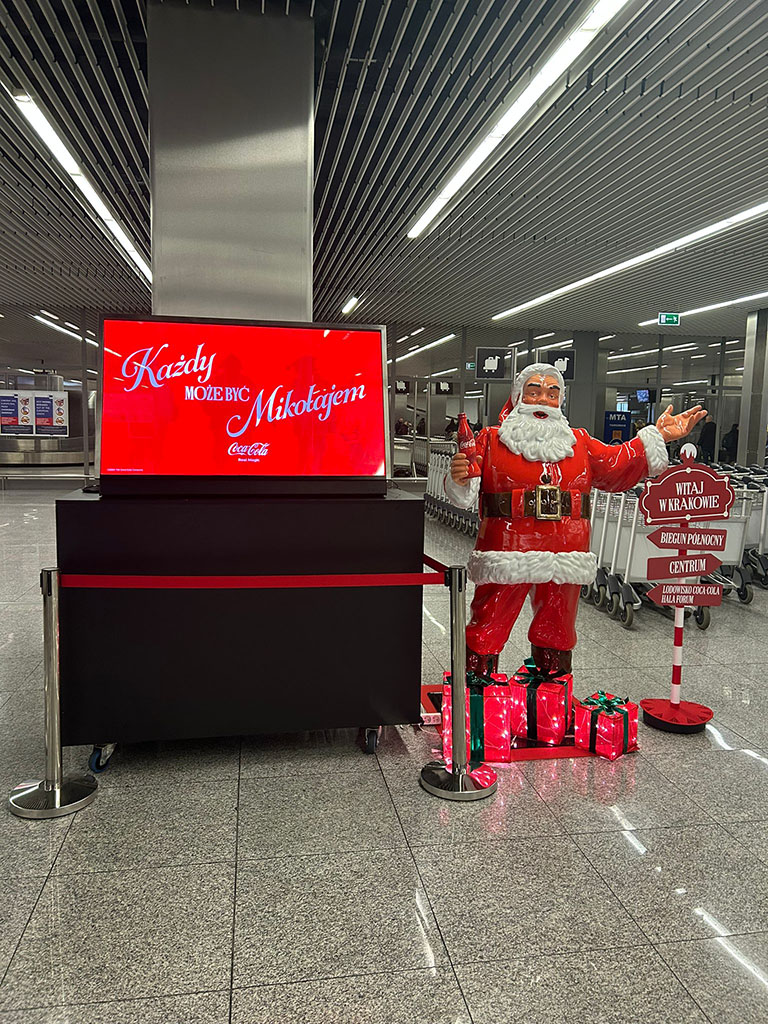 Anyone Can Be Santa: Coca-Cola's Christmas Campaign with Santa Figures 9 Figurka Świętego Mikołaja z butelką Coca-Coli i dekoracyjnymi prezentami przy informacji lotniskowej, obok stoi czerwony ekran z napisem