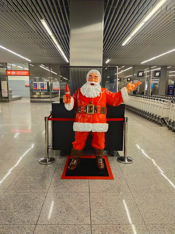 Anyone Can Be Santa: Coca-Cola's Christmas Campaign with Santa Figures 10 Figurka Mikołaja w czerwonym stroju z białym futrem stojąca na czarno-czerwonym podłożu, trzymająca czerwoną butelkę, w nowoczesnym hali z metalowymi sufitami i rzędem wózków po prawej stronie