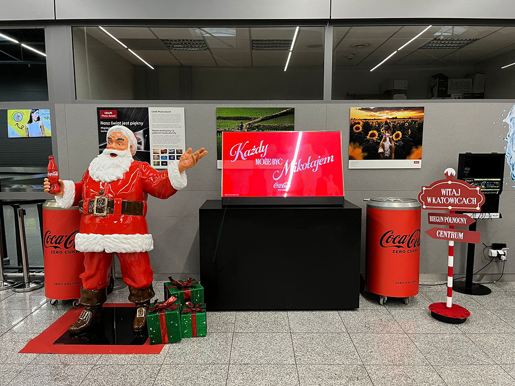 Anyone Can Be Santa: Coca-Cola's Christmas Campaign with Santa Figures 12 Figurka świętego Mikołaja w czerwonym ubraniu trzymająca butelkę Coca-Coli, stojąca obok dwóch zielonych prezentów z czerwonymi kokardami, na tle szarej ścianki z plakatami i czerwonych pojemników na napoje Coca-Cola Zero, oraz czerwony znak z napisami witającymi w Katowicach i wskazującymi kierunki.