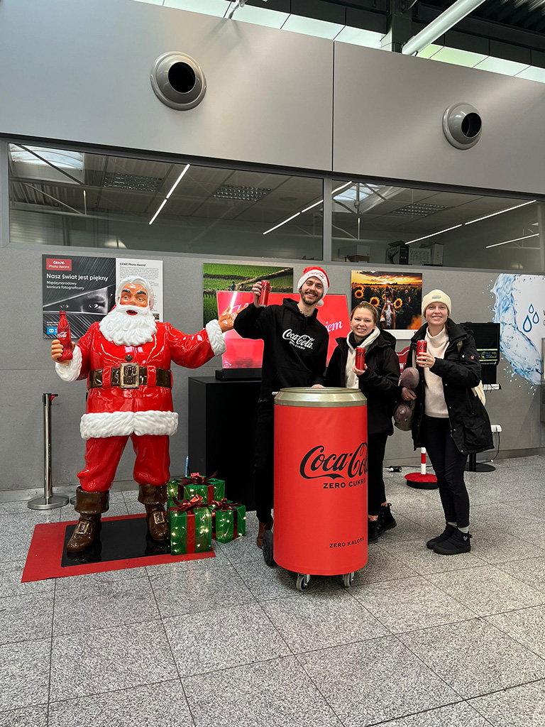 Anyone Can Be Santa: Coca-Cola's Christmas Campaign with Santa Figures 13 Trzy osoby stoją obok dużej figury Mikołaja w czerwonym stroju trzymającego butelkę Coca-Coli, każda z nich trzyma puszkę Coca-Coli, obok stoi duża puszka Coca-Coli na kółkach, tło to wnętrze budynku z szarymi ścianami i gablotami ze zdjęciami.