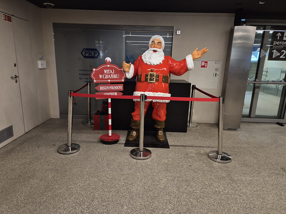 Anyone Can Be Santa: Coca-Cola's Christmas Campaign with Santa Figures 15 Figurka świętego Mikołaja w czerwonym stroju z białym futerkiem stojąca na dworcu w Gdańsku, obok czerwonego stojaka z napisem witającym w Gdańsku i kierunkami do Bieguna Północnego i Centrum, otoczona barierkami z czerwonym paskiem.