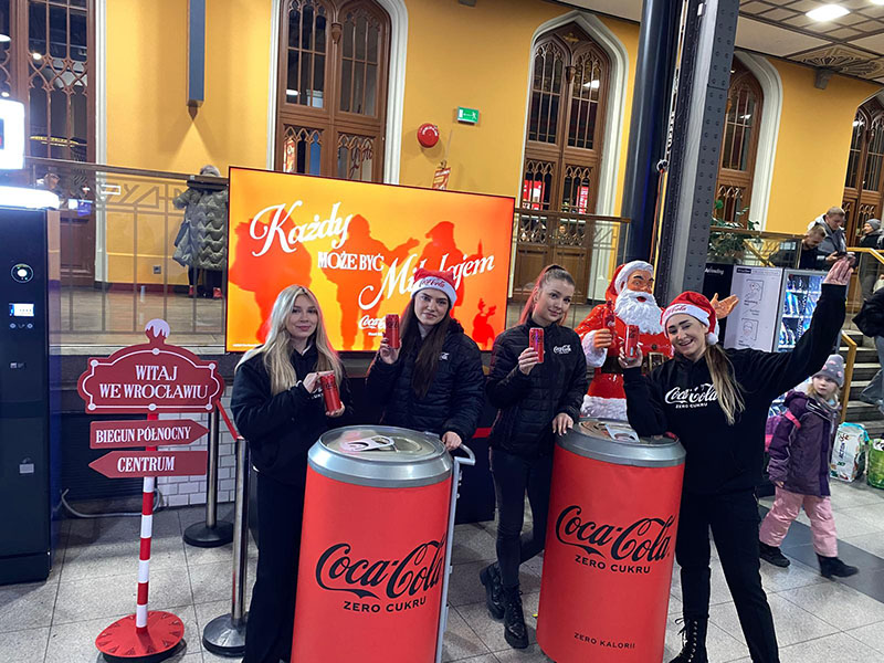 Anyone Can Be Santa: Coca-Cola's Christmas Campaign with Santa Figures 16 Cztery młode kobiety w ciemnych kurtkach i czapkach Mikołaja pozują z puszkami Coca-Coli przy dwóch dużych stojakach w kształcie puszek Coca-Coli Zero Cukru, na tle ekranu z napisem