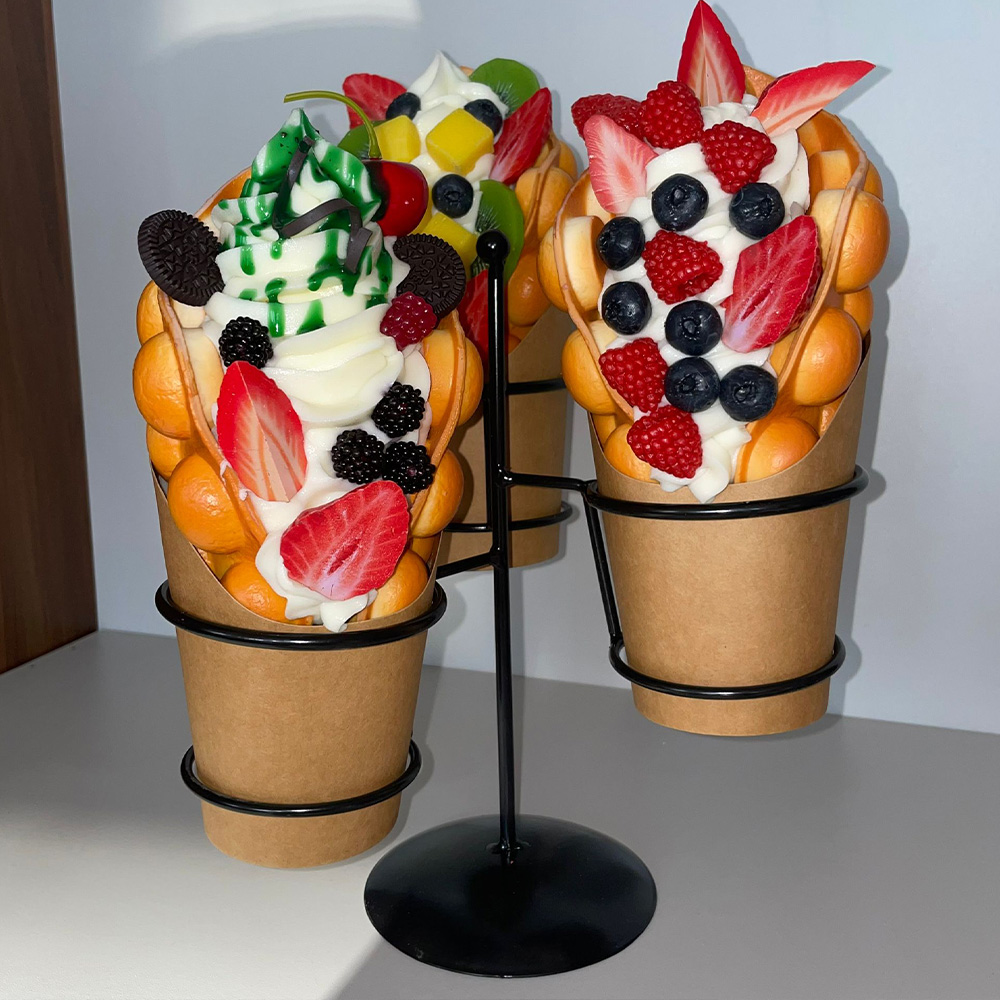Decorative Bubble Waffles in a Basket 2 Three bubble waffles filled with soft serve ice cream and assorted fresh fruits like strawberries, blueberries, raspberries, kiwi, mango, and topped with Oreo cookies, displayed on a black metal stand against a plain background.