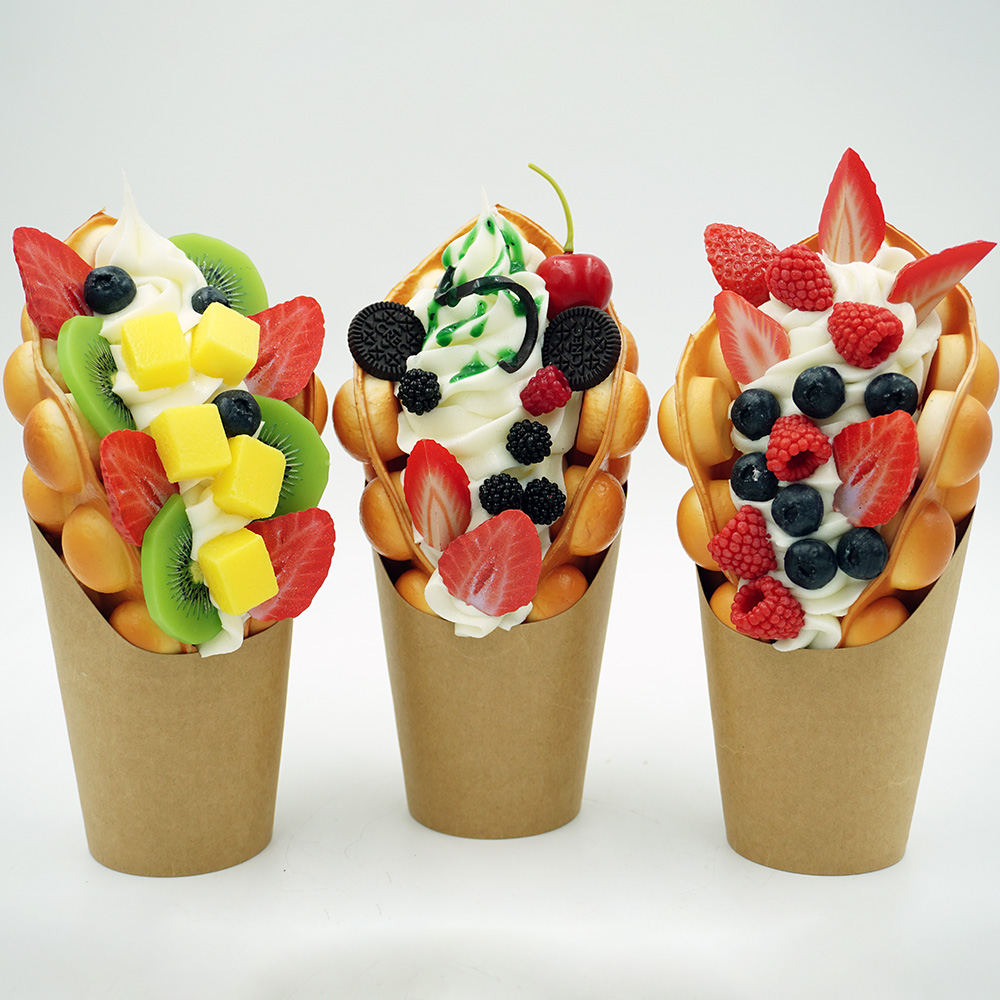 Decorative Ice Cream Basket – Motif I 6 Three bubble waffles in brown paper cups topped with whipped cream and various fruits including strawberries, blueberries, kiwis, mango cubes, raspberries, blackberries, a cherry, and mini chocolate cookies, arranged in a row on a white background.
