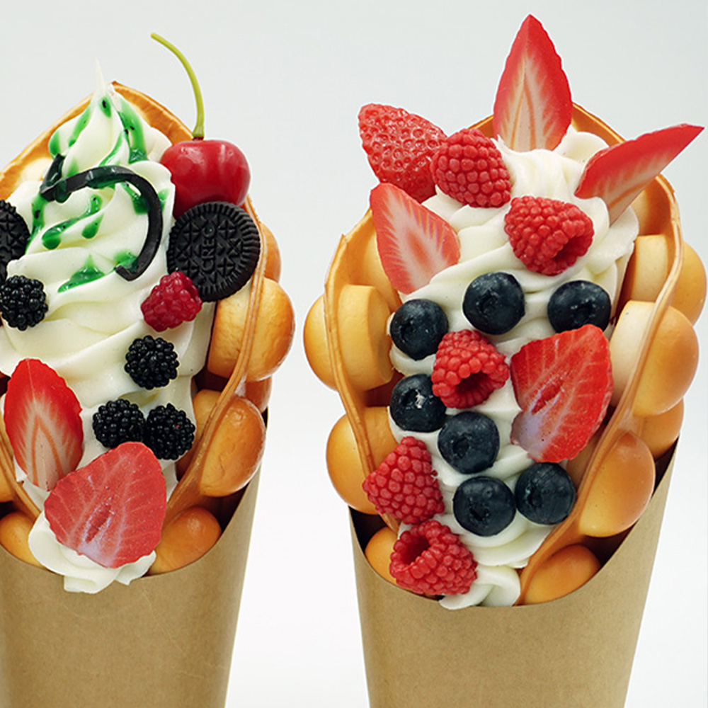 Decorative Bubble Waffles in a Basket 3 Two bubble waffle cones filled with soft serve ice cream topped with assorted berries, a cherry, mini Oreo cookies, and green syrup on a white background.