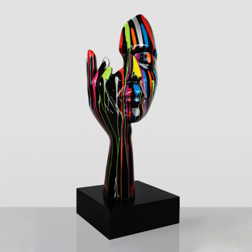 Glossy black sculpture of a hand holding a face, with vibrant multicolored paint streaks dripping down both the face and hand, mounted on a black square base against a plain gray background.