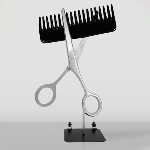 A metal sculpture depicting a pair of silver scissors holding a large black comb, mounted on a black rectangular base against a plain light gray background.